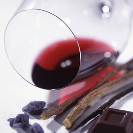 A Glass Of Red Wine Lying On Its Side, With Candied Violets, Dark Chocolate, A Vanilla Pod And Liquorice Roots by Blueberrystudio