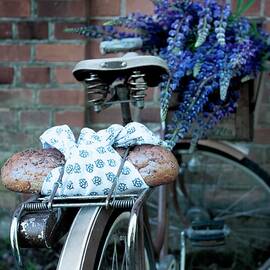 Bread Wrapped In Blue And White Cloth On Bicycle Luggage Rack With Bouquet Of Lupins In Crate On Handlebars In Front Of Brick Wall by Annette Nordstrom