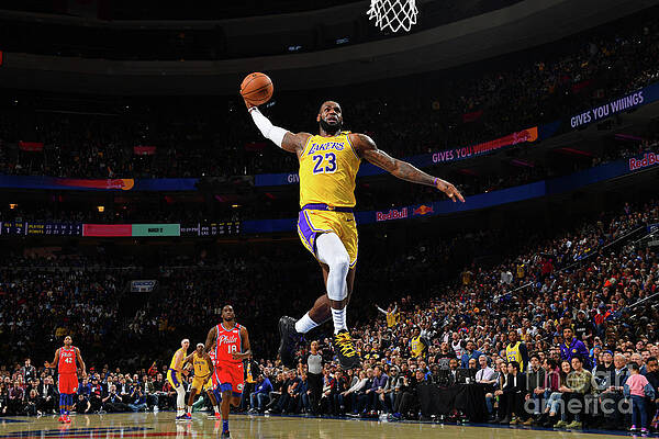LeBron James (#23) of the Los Angeles Lakers performs a dynamic one-handed dunk during an NBA game. He is captured mid-air in his iconic yellow Lakers jersey against a red opposing team (appears to be Chicago Bulls based on uniforms). The arena is packed with fans in the background, creating an energetic atmosphere. The image showcases James's athleticism and vertical prowess. The photo is taken from a low angle, emphasizing the height and power of the play. Professional arena lighting and signage are visible, and other players can be seen on the court below. The mood is triumphant and action-oriented.